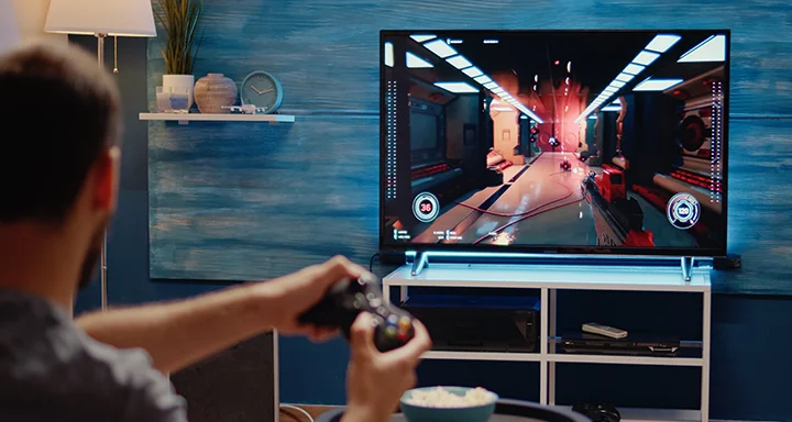Elinde oyun kumandası ile bir oyuncunun oyun oynarken omzunun üzerinden bakılan görüntüsü, arka planda bir TV monitörü.