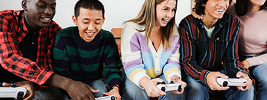 Пятеро юных геймеров сидят дома на диване, четверо держат в руках контроллеры PS5 и играют вместе.