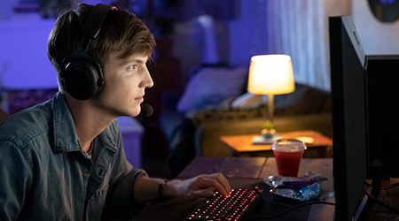 A young man looking at a PC monitor in front of a backlit keyboard with a lamp in the background
