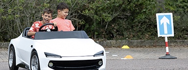 Two smiling kids driving a Young Driver’s white miniature practice car