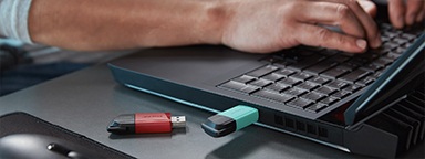 2 USB DT Exodia M, uno con un cappuccio verde e l'altro rosso, sul di una scrivania con una persona che lavora a un laptop sullo sfondo