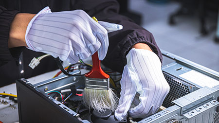 A technician wearing gloves uses a brush to clean the inside of a computer case on a workbench.