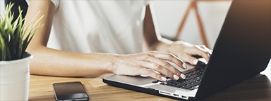 Gambar close-up seorang wanita muda sedang menulis di keyboard laptopnya dengan tanaman dan telepon di sebelahnya di atas meja