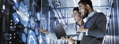 Immagine di uno specialista IT che tiene in mano un laptop mentre parla con un tecnico server in una sala server di un data center, con le icone del cloud e di uno scudo raffigurate sui rack server