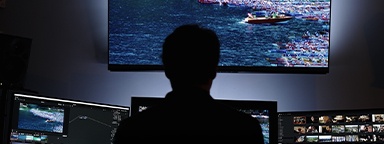 A silhouette of a video editor working with monitors and a larger screen displaying his project in a dimly lit room