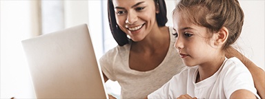 una madre y su hija sonriendo sentadas en una mesa usando una computadora portátil en casa
