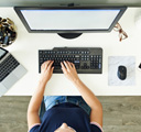 Vista superior de las manos de una persona que trabaja con un teclado en un escritorio con un monitor, computadora portátil, mouse y teléfono móvil cerca