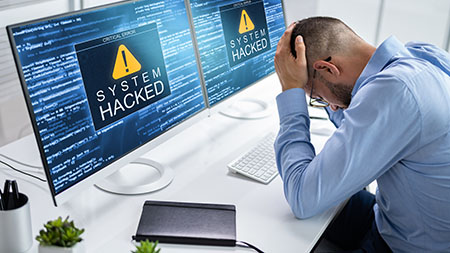 A defeated office worker cradles his head in his hands as he sits in front of two monitors displaying the error message ‘System Hacked’.