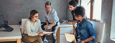 Four office workers collaborate using various devices.