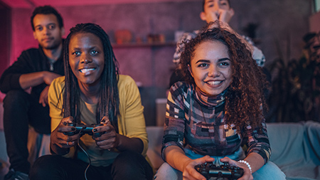 Dwie osoby na kanapie, skupione na grze, trzymają kontrolery PlayStation. Za nimi stoją dwie kolejne osoby, które im kibicują.