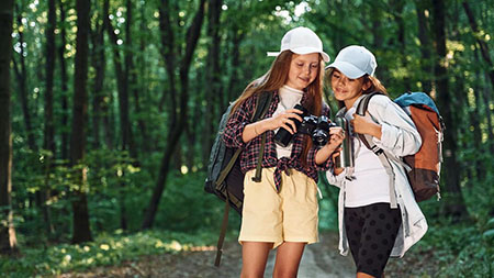 Two young girls on a forest path with hiking backpacks share a look at their camera screen.