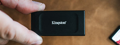 A hand holds a Kingston XS1000 external SSD over brown background.