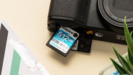 桌上放着一台照相机，一张存储容量为 512GB 的 Kingston Canvas Go!Plus 存储卡已经插入一半。桌上还放着一盆多肉植物和一个文件夹。