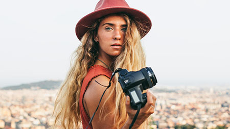 Une jeune femme debout devant une vue de la ville tient un appareil photo, l’offrant au spectateur.