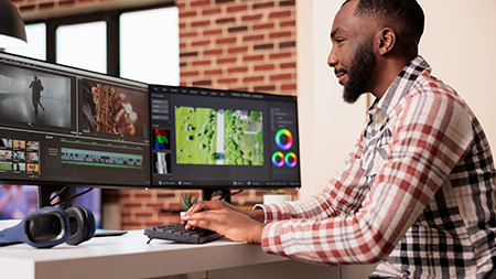 Man sits at a two-monitor computer desk, edits video footage via keyboard