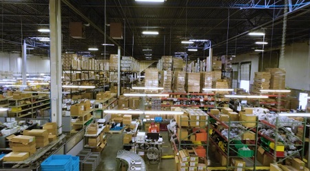 Wide shot of a full and well-organised warehouse space full of racks of boxes and trays, one employee behind a metal desk working at a computer