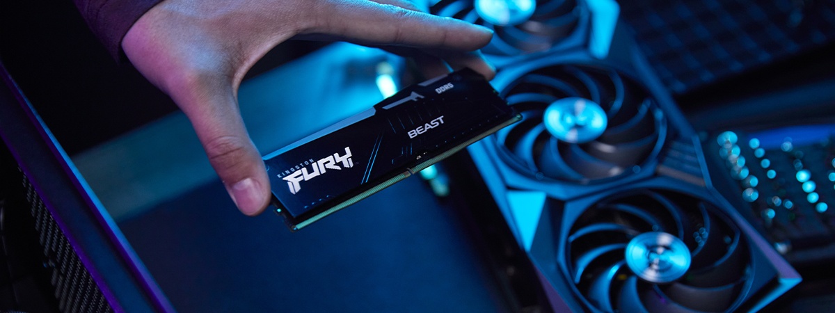 Primer plano de una mano sujetando un m&oacute;dulo de memoria Beast DDR5 RGB con piezas de PC y un ventilador desenfocados sobre la mesa