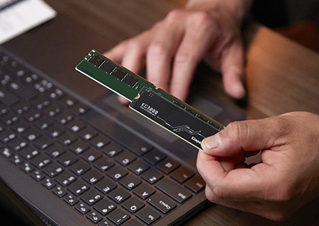 Kingston KC3000 SSD と DDR5 メモリモジュールを持つ手と、その後ろにある机の上に置かれたノートパソコンのクローズアップ。