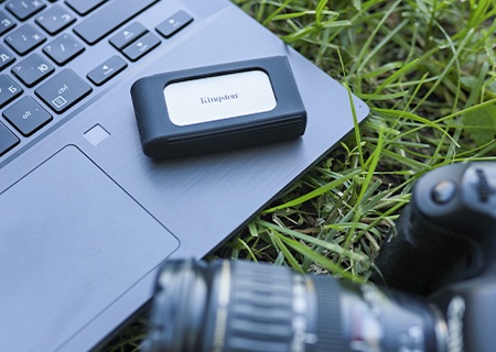 Ổ SSD XS2000 trên một chiếc máy tính xách tay trên bãi cỏ, một chiếc camera nằm ở tiền cảnh