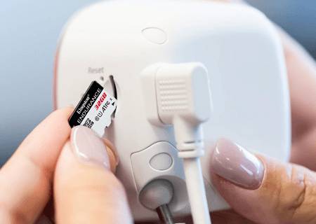 The rear side of a white security camera with a microSD card being inserted by fingers