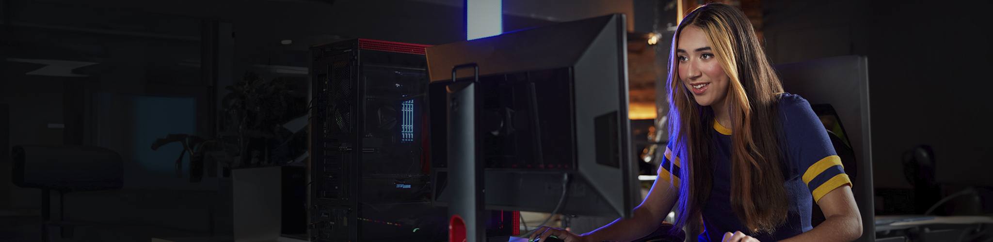 A young woman gamer sits at a desk with a gaming PC and GRB keyboard, she is looking at a flat screen monitor.