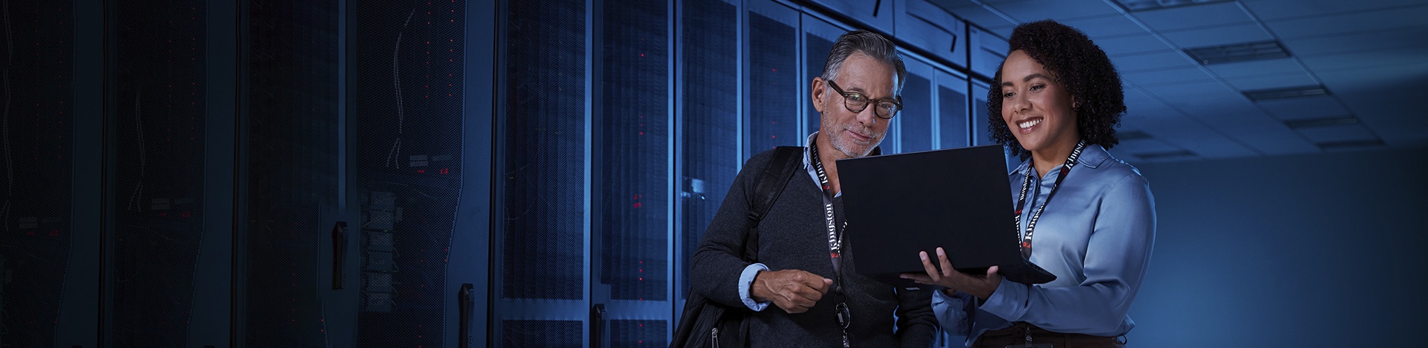 Two IT workers stand in a dimly lit server room looking at a laptop