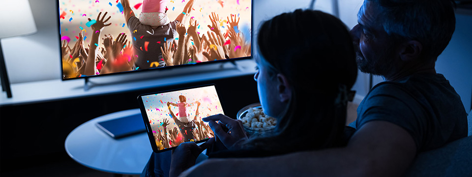 Organizing Your Media Collection A family in a dark living room look at a tablet and television both displaying a child on a parent’s shoulders at a crowded parade.