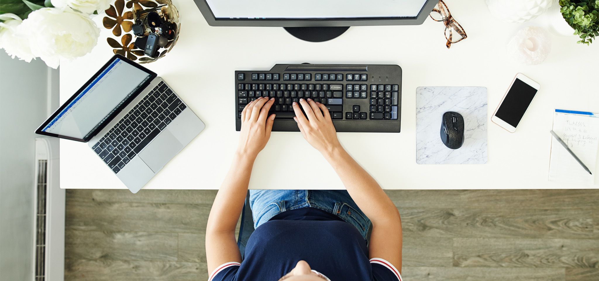 Widok od góry rąk osoby pracującej na klawiaturze przy biurku komputerowym, z monitorem, laptopem, myszą i smartfonem w pobliżu