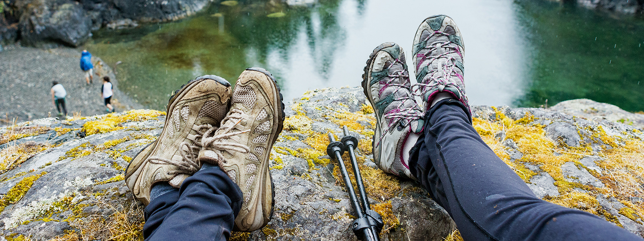 Os pés de duas pessoas pendurados sobre a beira de uma rocha com vista para um lago.