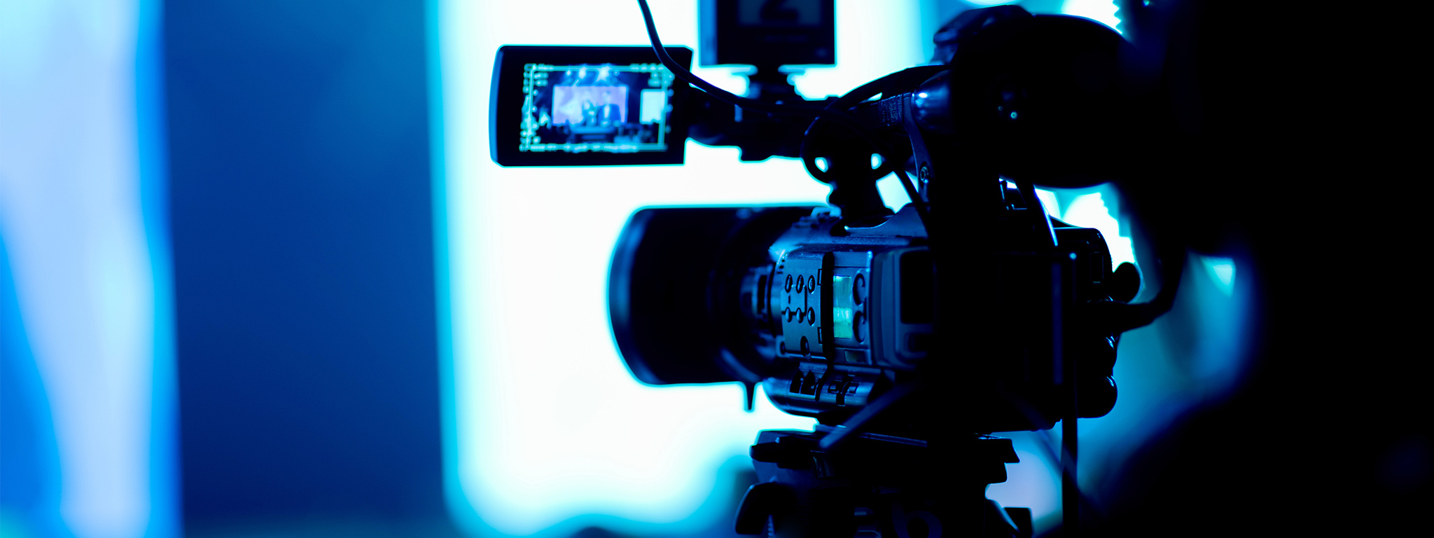 A person in soft focus operates a camera in a dark room with various peripherals attached, a display showing on a fold out arm.