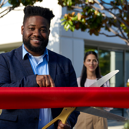 A young professional man uses a large pair of novelty scissors to cut a red ribbon.