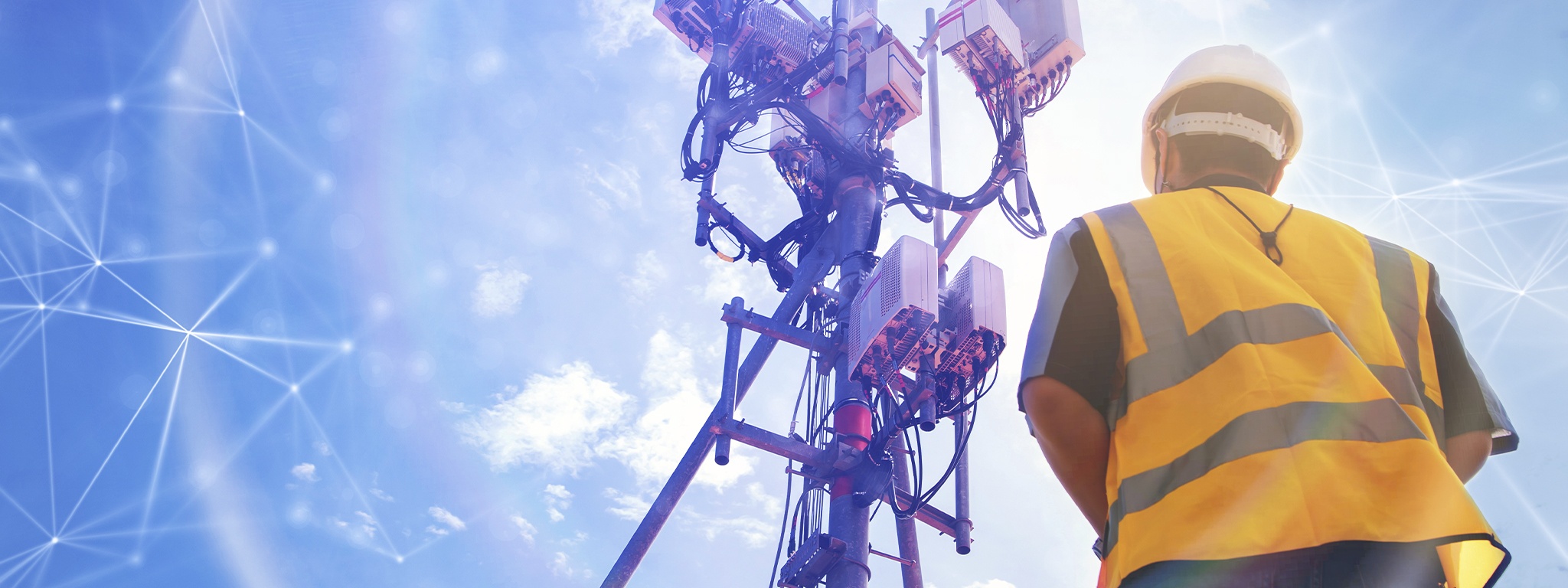tampak belakang seorang teknisi berhelm yang bekerja di lapangan dengan menara telekomunikasi di hadapannya