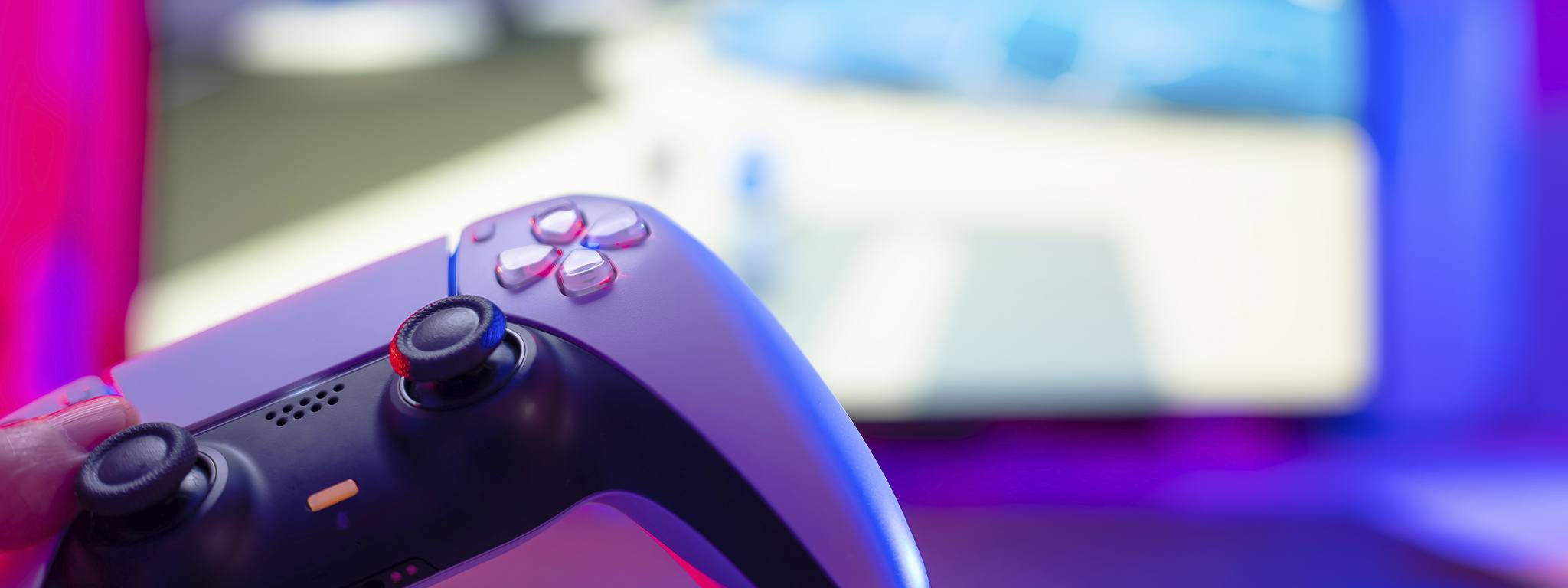 A close-up shot of a PlayStation 5 controller with a blurry background of a game on the TV