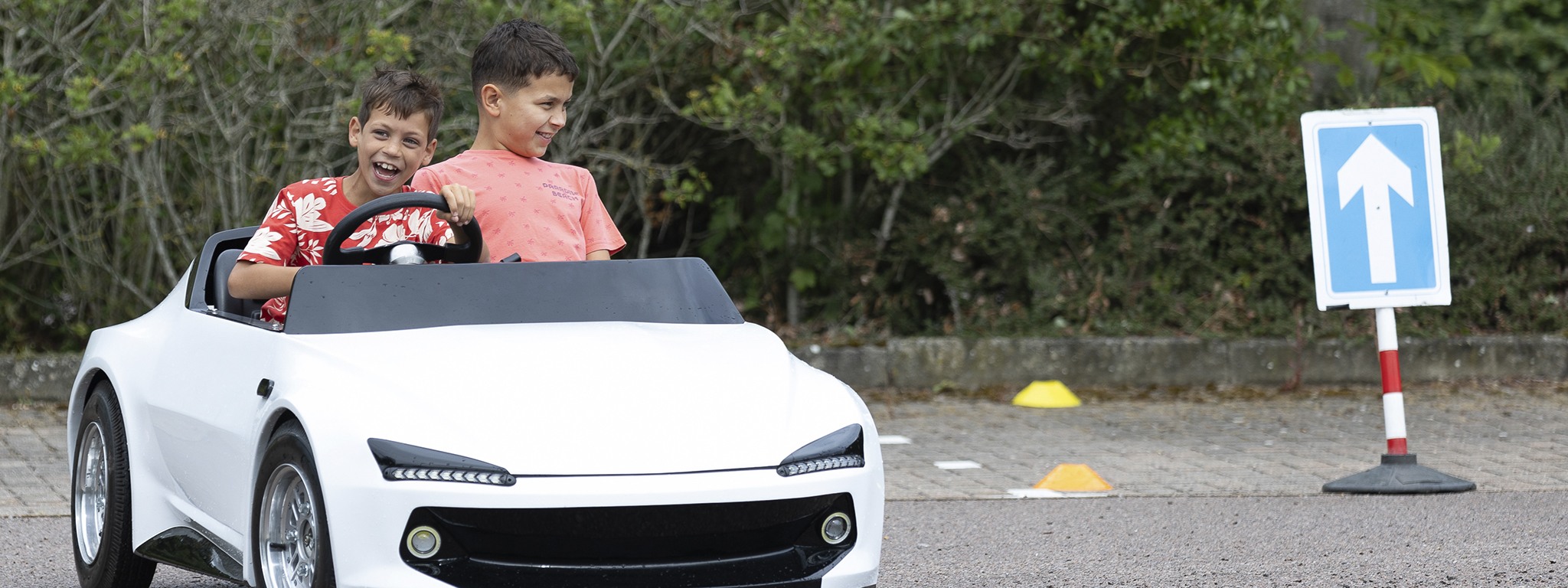 2 çocuk gülümseyerek Young Driver'ın beyaz minyatür alıştırma arabasını sürüyor