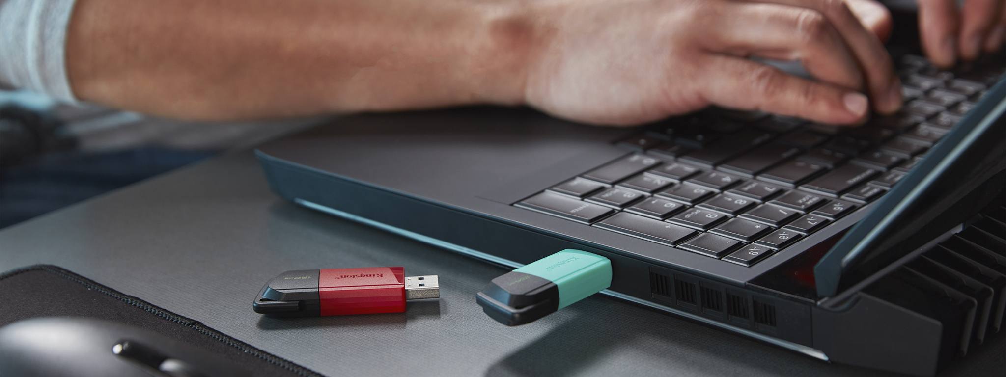 Hai ổ USB Flash DT Exodia M, một có nắp xanh lá và một có nắp đỏ, đặt ở cạnh bàn, phía hậu cảnh là một người đang sử dụng máy tính xách tay
