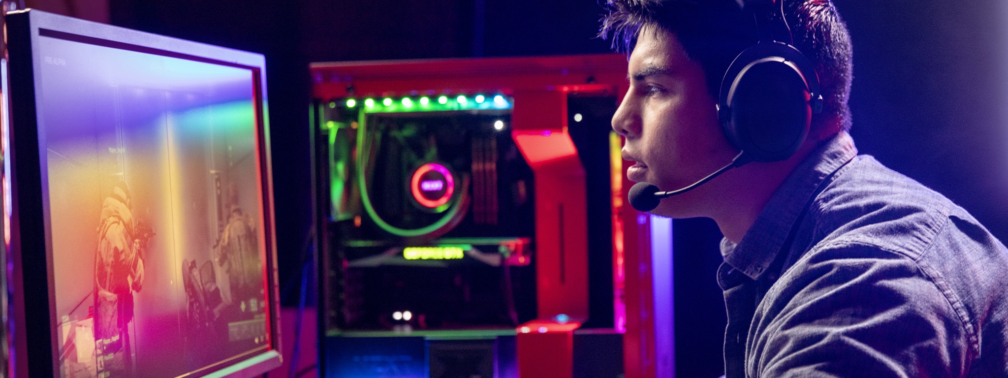 a young gamer sitting on his desk wearing a headset, playing PC FPS games with his computer desktop in the background