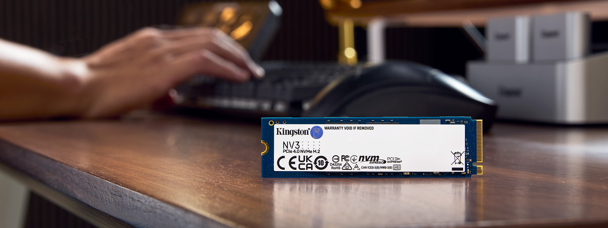 Một ổ SSD NV3 NVMe M.2 đặt thẳng đứng trên cạnh bàn, ở hậu cảnh là đôi bàn tay đặt trên bàn phím.