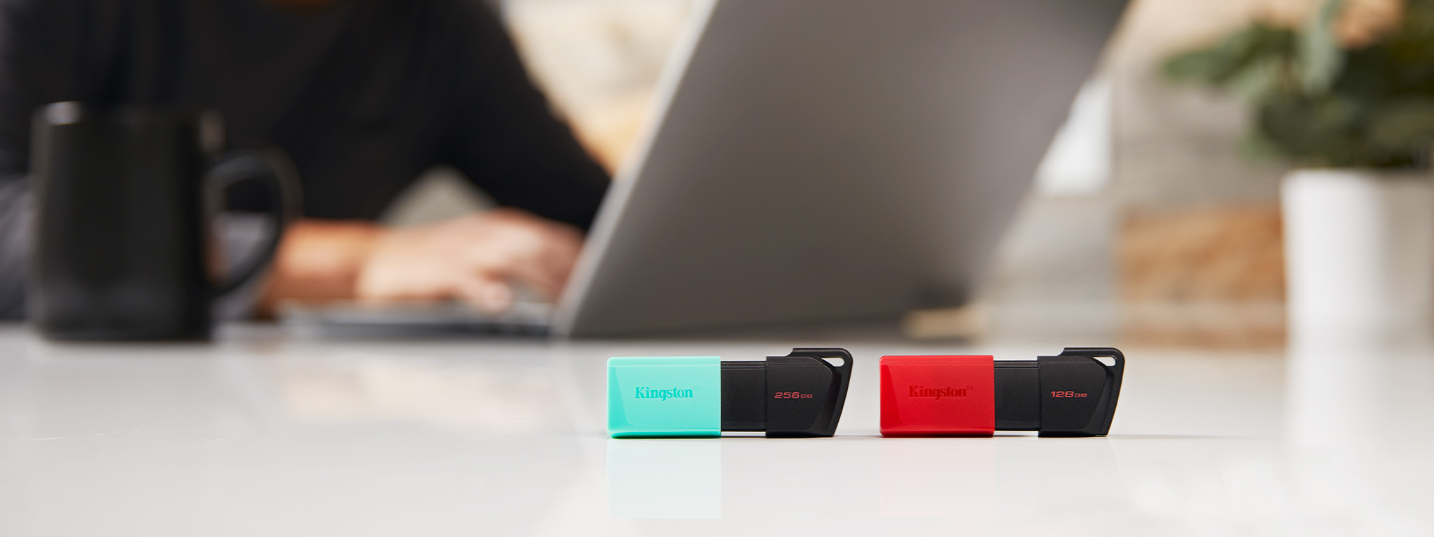 Dwie pamięci flash USB DT Exodia M, jedna z zieloną nasadką, druga z czerwoną, leżące na krawędzi biurka, w tle osoba korzystająca z laptopa