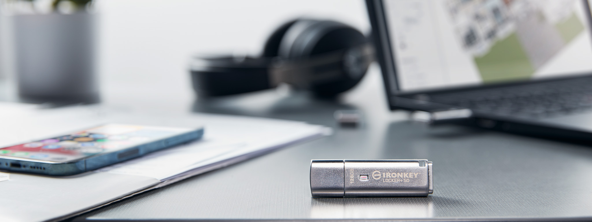 An IronKey Locker+ 50 sits on a desk with a laptop and headset in the background