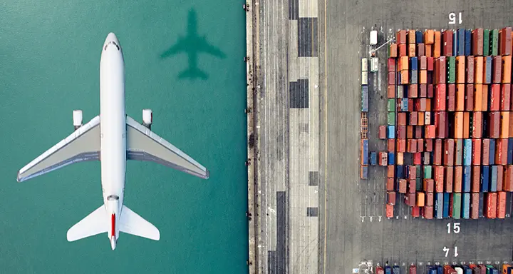 An overhead view of a plane flying perpendicular to multicolored containers on a dock. 