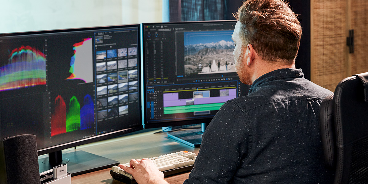 Um profissional de tecnologia trabalhando em um vídeo em uma mesa com dois monitores, em frente a uma janela com várias plantas interiores.