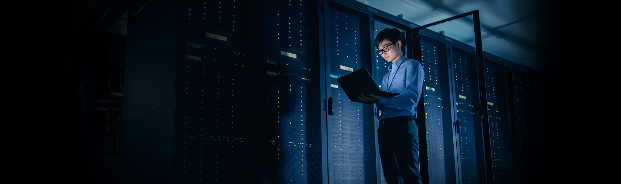 A professional standing in front of a bank of servers, holding and using a laptop. 