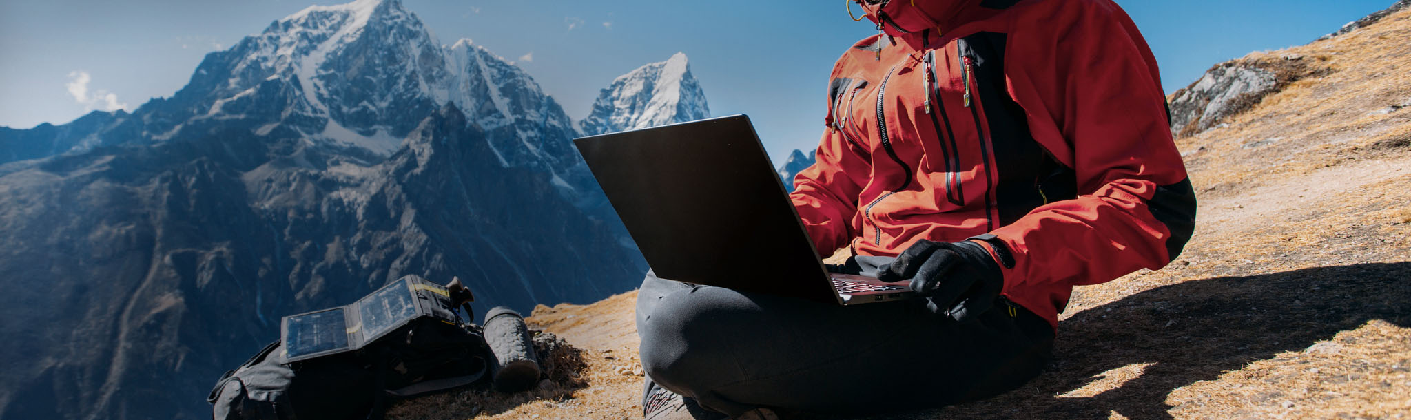 A hiker sitting on a mountain slope using a laptop.]